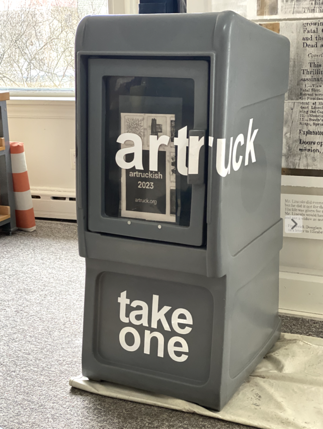 a photo of a free newspaper box with the words artruck and take one on the front. the box is grey and the letters are white. inside of the box are tall newsprint papers with images of all of the artwork from the artruckish 2023 exhibition.